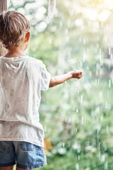 Blond little boy in white t-shirt holds hand under falling down rain drops standing on country house terrace backside view