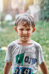 Disgusted little boy in wet t-shirt stands under warm heavy summer rain falling down on lush green...