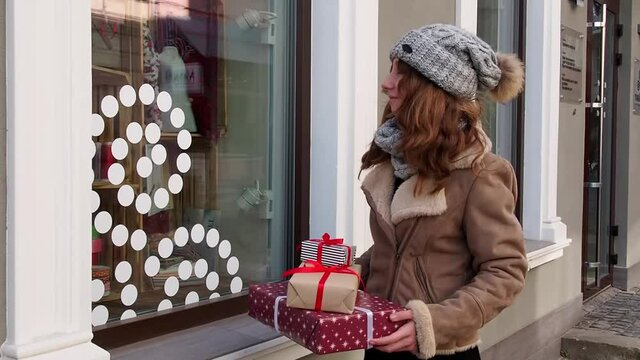 Teengirl in grey hat, scarf and brown jacket holding a set of christmas gifts near shops window. Happy young woman preparing holidays presents. Concept of shopping, giving and receiving gifts.