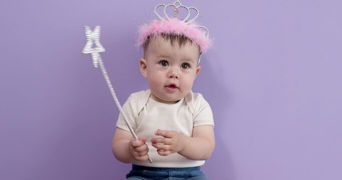 Beautiful little baby girl holding magic wand stick in hands, studying it, isolated on purple background. Toddler child in casual wear is looking cute, excited girl in crown posing. Childhood concept