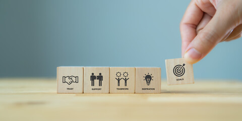 Business goals achievement concept with collaboration and teamwork. Businessman holds the wooden cube with symbols ; goals,  trust, support, teamwork and inspiration on grey background and copy space.