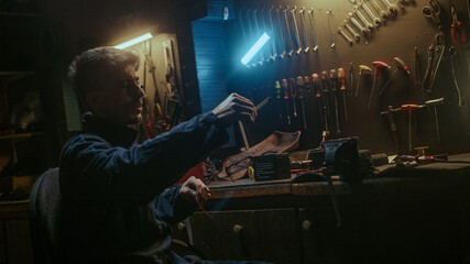 Young blacksmith male with workwear sitting on chair, taking measurements and working on workbench in his workshop