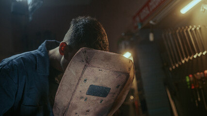 Young blacksmith man with welding shield using welding machine for welding metal construction on...