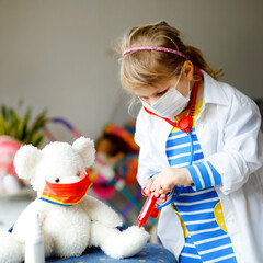 Little girl makes injection to teddy bear. Cute child with medical mask playing doctor, holding...