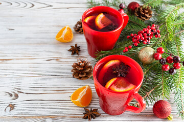 Mulled wine with orange, apples, cinnamon and star anise in red ceramic cups on a light wooden table. Fir branches with red berries and cones in the background. Place for text