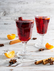 Two transparent glass wine glasses with fragrant mulled wine from red wine with orange on a light wooden table. Pieces of tangerines, cinnamon tubes in the background