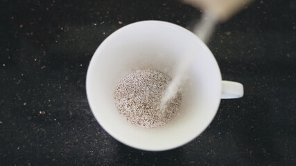 Top view of a white mug on a black table in which instant coffee with milk is poured. A beautiful shot of making fast-brewed coffee from bags in the morning in slow motion. 4K.
