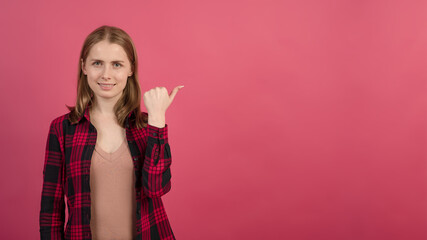 Young and attractive caucasian blonde teenager girl pointing her thumb to the advertising space, for copy space on a pink studio background.