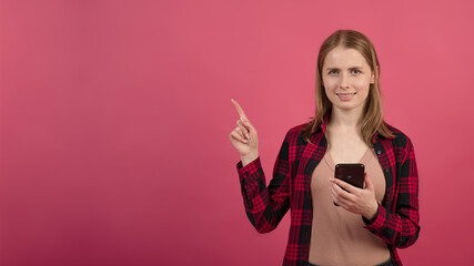 A young and attractive blonde Caucasian girl in a shirt using a mobile phone and pointing her finger at an empty advertising space on a pink studio background.