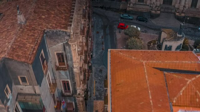 An aerial top-down view of a narrow historical street in Catania, Sicily. Traditional European buildings with orange tiled rooftops frame the ancient alley below.