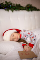 Merry Christmas and Happy Holidays. The little happy girl sleep on the bed wrote a letter to Santa Claus and holds the envelope in her hand.