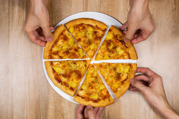 Top view of hands taking slices of delicious and crispy hawaiian pizza. Group of hungry friends sharing delicious lunch on wooden table.