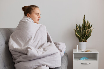 Sad tired woman with hair bun sitting on sofa wrapped in blanket felling very bed, having covid...