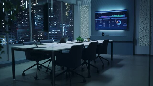 Modern Empty Meeting Room with Big Conference Table with Various Documents and Laptop on it. Wall TV Showing Company's Growth, Statistics, Graphs and Pie Charts. Late Evening or Night City Outside.