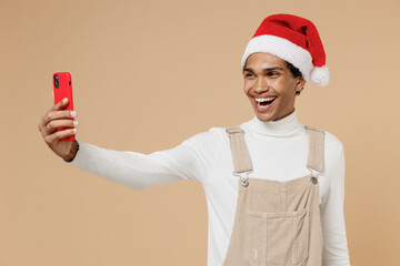 Young happy african man 20s wear Santa Claus red Christmas hat doing selfie shot on mobile cell phone isolated on plain pastel beige background studio portrait Happy New Year 2022 celebration concept