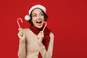 Young smiling amazed woman 20s wear Santa Claus Christmas red hat hold candy cane stick lollipop look aside isolated on plain red background studio portrait. Happy New Year 2022 celebration concept.