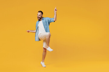 Full body young overjoyed excited caucasian man in blue shirt do winner gesture with raised up leg clench fist celebrate isolated on plain yellow background studio portrait. People lifestyle concept.