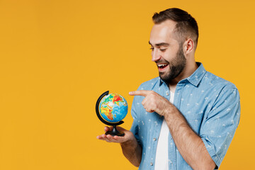 Young smiling happy cheerful friendly caucasian man 20s in blue shirt white t-shirt hold in hands spin Earth world globe isolated on plain yellow background studio portrait. People lifestyle concept.