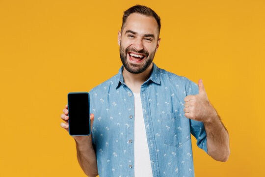 Young smiling happy man 20s in blue shirt white t-shirt hold in hand use mobile cell phone with blank screen workspace area show thump up gesture isolated on plain yellow background studio portrait.