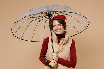 Young smiling happy woman 20s wears red turtleneck vest beret hold transparent umbrella look camera isolated on plain pastel beige background studio portrait. People lifestyle rainy weather concept. - obrazy, fototapety, plakaty