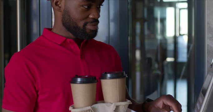 Courier african man doing fast food delivery, dials the house number in intercom. Bearded male in red uniformv came to customers, holding cup of coffee in hands,very pleasant and nice