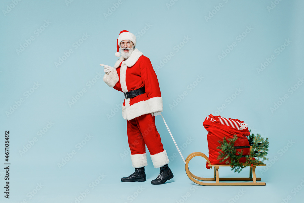 Wall mural Full body old Santa Claus man in Christmas hat red suit clothes go with gifts sled point finger aside on workspace isolated on plain blue background studio. Happy New Year 2022 merry ho x-mas concept.