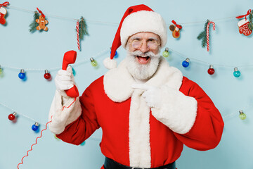 Old Santa Claus man 50s wear Christmas hat red suit talk speak point finger on handset phone isolated on plain blue background studio. Happy New Year 2022 celebration merry ho x-mas holiday concept.