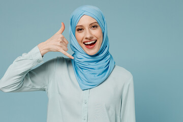 Young smiling arabian asian muslim woman in abaya hijab doing phone gesture like says call me back isolated on plain blue background studio portrait. People uae middle eastern islam religious concept.