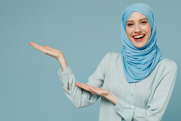 Young arabian asian muslim woman in abaya hijab point hands arms aside copy space workspace area isolated on plain blue background studio portrait. People uae middle eastern islam religious concept. - obrazy, fototapety, plakaty