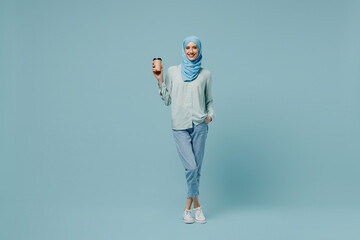 Full body young arabian asian muslim woman in abaya hijab hold takeaway delivery craft paper brown cup coffee to go isolated on plain blue background People uae middle eastern islam religious concept