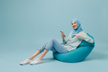 Full body young arabian asian muslim woman in abaya hijab sit in bag chair hold point on mobile...