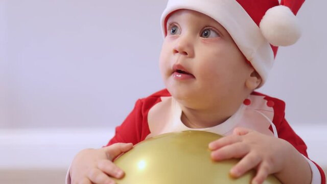 portrait of cute caucasian infant baby kid in Santa costume red hat playing with golden balloon, charming toddler trying to climb the ball looking to camera smiling opening mouth with drooling. x-mas