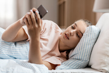 people, bedtime and rest concept - happy smiling girl with smartphone lying in bed at home