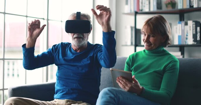Laughing mature woman sitting on sofa with tablet while laughing at senior father in VR goggles being excited with new impressions of virtual reality