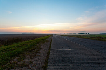 An empty asphalt road through the fields and forest in a thick fog at sunrise