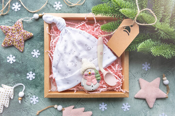 Gift box with a pink paper filler with baby hat, spoon and toy snowmen on a green background with Christmas decor. New Year's gift for the birth of a baby. Cozy christmas concept.