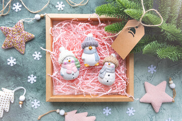 Gift box with toys on a green background with Christmas decor. There are three ceramic toys in a box with a pink paper filler, snowmen and a penguin in knitted hats and scarves. Cozy christmas concept