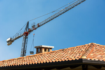 Naklejka premium Construction site with tower crane and a closeup of a roof with terracotta tiles (Coppo in Italian language) and gutter on a clear blue sky. Construction industry concept. Italy, Europe. 