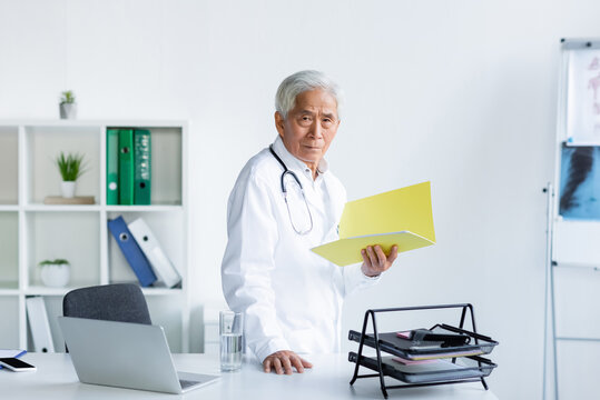 Senior asian doctor holding paper folder near devices and glass of water in clinic