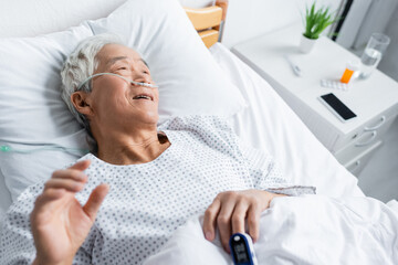 High angle view of smiling asian patient with pulse oximeter and nasal cannula lying on bed in clinic