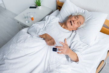 High angle view of sick asian patient with pulse oximeter lying on bed near pills in clinic