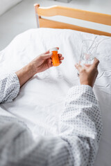 Cropped view of senior patient holding water and pills on bed in clinic