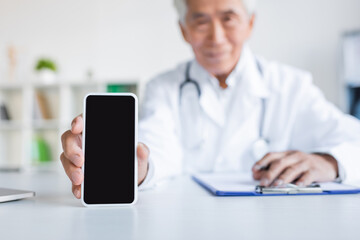 Blurred asian doctor holding smartphone with blank screen near clipboard in hospital