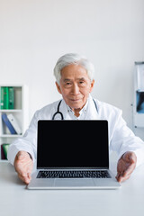 Fototapeta na wymiar Elderly asian doctor looking at camera near laptop with blank screen in hospital