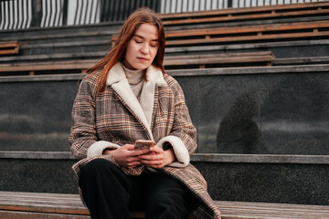 Young woman in coat with smartphone in hands sitting alone on wooden bench on a street. Working on mobile device out of home outdoors. Female freelancer, blogger. Texting, chatting, reading.