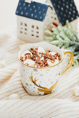 Cup of hot chocolate with marshmallows on white knitted plaid. Ceramic little houses and fir branch on background. Christmas, winter holidays in cozy home concept.