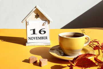 Calendar for November 16: a cup of tea with branches of girlish grapes against the background of a decorative house with the name of the month in English and the number 15, gray background