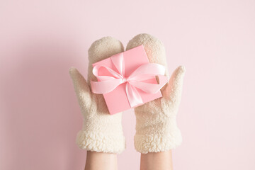 Female hands in mittens holds  Christmas pink gift with satin bow on pink background. Hands with gift. Flat lay, top view, copy space