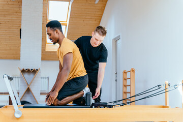 Fitness instructor corrects and controls the Pilates exercise that his african american male student is doing on Reformer bed in health center, gym with modern interior.