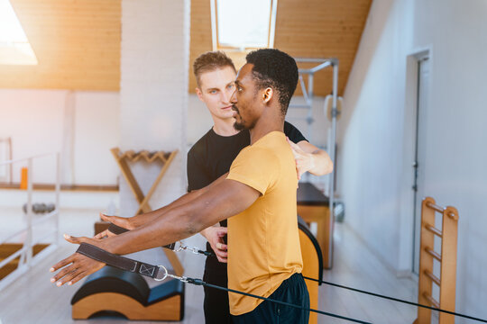 Fitness instructor corrects and controls the Pilates exercise that his african american male student is doing on Reformer bed in health center, gym with modern interior.